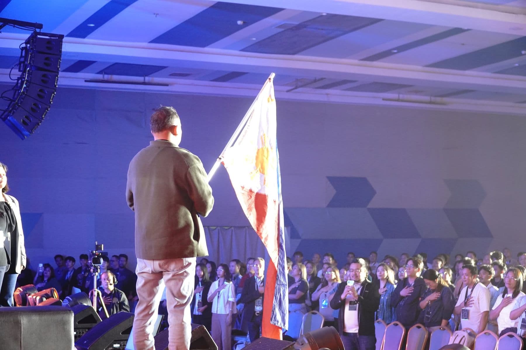 A man holding the Philippine flag on stage as delegates stand with hands on their hearts