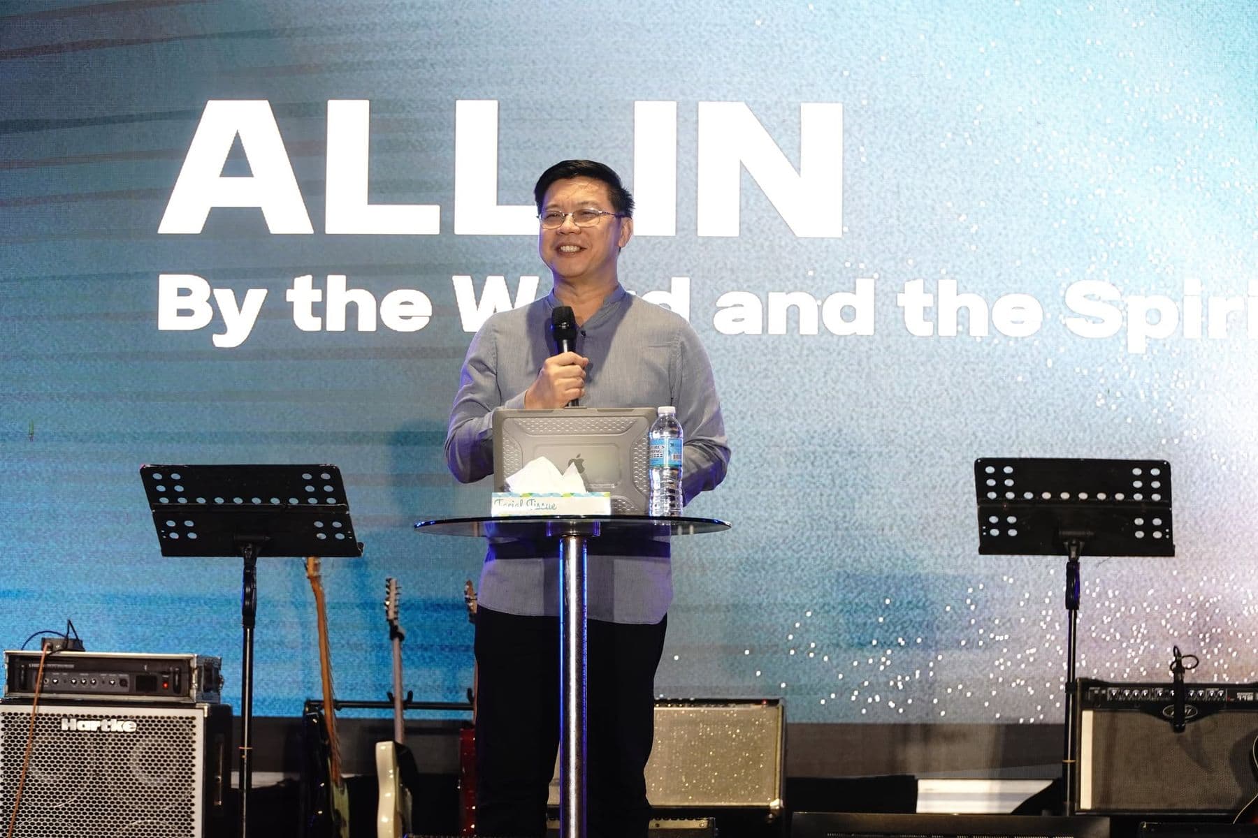 A speaker teaching at a lectern beneath the words All In, By the Word and the Spirit