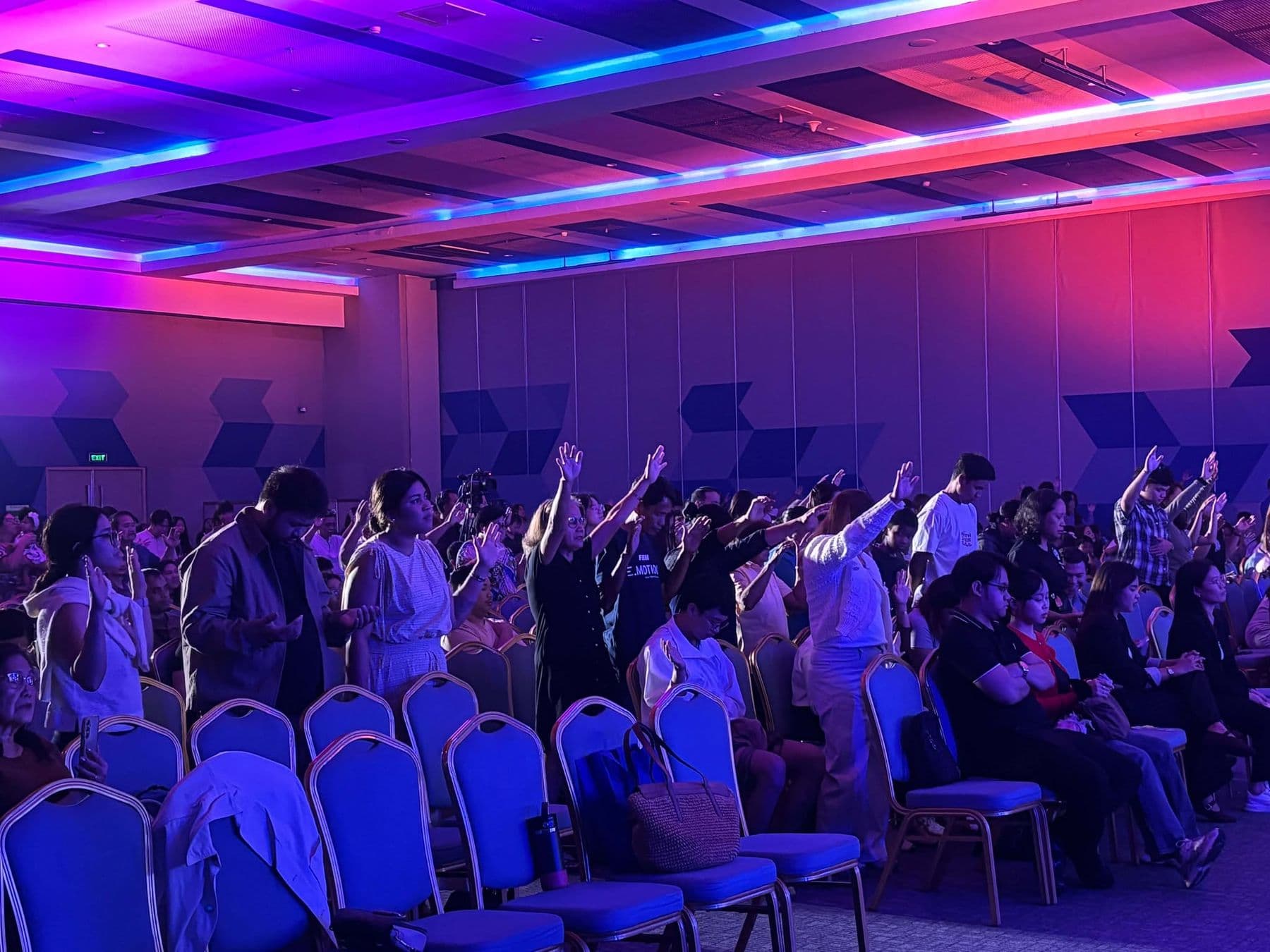 Rows of delegates worshipping with arms lifted under purple light